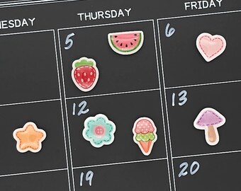 May include: A black calendar with white grid lines and handwritten numbers. Colourful, illustrated stickers of a strawberry, watermelon, flower, ice cream cone, star, heart, and mushroom are placed on the calendar.