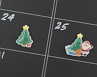 May include: A black calendar with the numbers 24 and 25. The calendar has Christmas-themed stickers, including a green Christmas tree and a Santa Claus figure. The stickers are placed on the calendar.