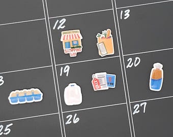 May include: A black calendar featuring white numbers and various stickers. Stickers depict a grocery store, shopping bag, eggs, a receipt with a sale tag, a white bag, and a bottle with blue liquid.