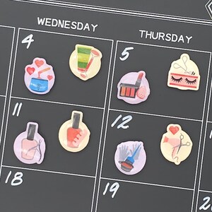 May include: A black calendar with white grid lines and the days of the week. The calendar features colorful stickers with beauty-related illustrations, including nail polish, lipstick, and a hairdryer. The word "Wednesday" and "Thursday" are visible at the top.