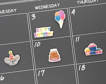 May include: A black calendar with white grid lines and numbers. Birthday-themed stickers are placed on the calendar, including a koala with a party hat, a cake, balloons, gifts, and the words "Happy Birthday."