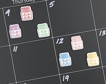 May include: A black calendar with white numbers and colourful birthday cake stickers. The stickers are on the days 4, 11, 12, 18, and 19. The cake stickers are red, purple, green, blue, and yellow.