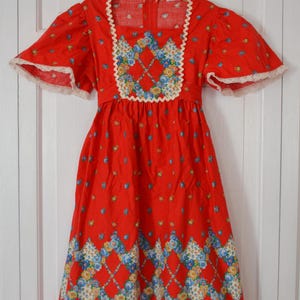 May include: A vintage red dress with short sleeves and a square neckline. The dress features a floral pattern and white lace trim on the sleeves and neckline. The dress is hanging on a wooden hanger.