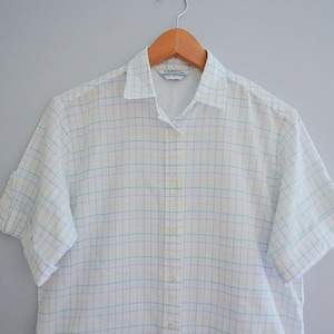 May include: A white button-down shirt with a plaid pattern in light blue and yellow. The short-sleeved shirt is hanging on a wooden hanger. The shirt has a collar and buttons down the front.