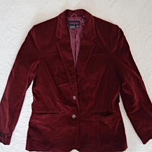 Peut inclure: Un blazer en velours bordeaux avec un revers cranté, deux poches avant et une fermeture à deux boutons. La veste est posée à plat sur une surface blanche à motifs. Le blazer a des manches longues et une coupe ajustée.