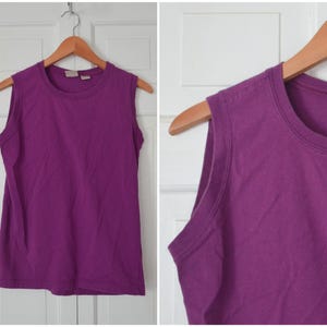 May include: A sleeveless, solid purple tank top hanging on a wooden hanger. The top has a crew neck and is made of a soft, likely cotton, material. The tank top is a vibrant shade of purple.