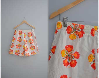 May include: White shorts with a vibrant floral pattern in shades of orange and yellow. The shorts are hanging on a wooden hanger against a gray background. The elastic waistband is visible.