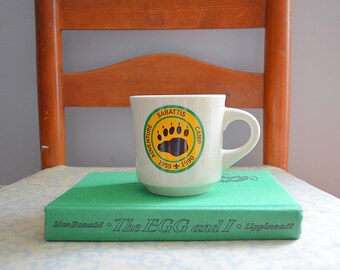 Puede incluir: Una taza de cerámica blanca con un logotipo de pata de oso y el texto "SABATTIS ADVENTURE CAMP 1959-1990" se encuentra sobre un libro verde titulado "The EGG and I". La taza tiene un asa curvada.