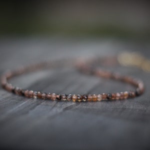 May include: A delicate brown and clear gemstone necklace with a simple design. The necklace is made of small, round beads and is strung on a thin chain.