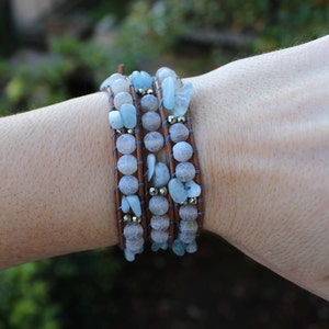 Puede incluir: Un brazalete de cuero de múltiples hebras con cuentas de piedras preciosas de color azul claro y gris. El brazalete está envuelto alrededor de la muñeca de una persona.