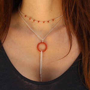 Puede incluir: Un collar de plata con un colgante circular hecho de cuentas de cornalina naranja. El colgante tiene un borla de cadenas de plata.