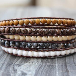 Puede incluir: Un brazalete de múltiples hilos con cordón de cuero marrón y diversas cuentas de piedras preciosas. Las cuentas son en tonos de marrón, blanco y gris.