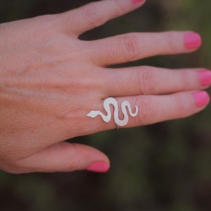 Op de afbeelding: Een zilveren slangenring met een minimalistisch ontwerp. De ring wordt gedragen op een vinger met roze nagellak.