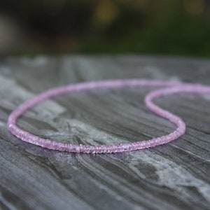 May include: A delicate pink gemstone necklace made of small, faceted beads. The necklace is laid out on a textured, gray surface.