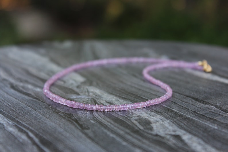 May include: A delicate pink gemstone necklace made of small, faceted beads. The necklace is laid out on a textured, gray surface.