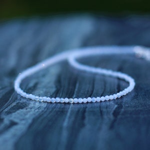 Può includere: Una collana delicata realizzata con piccole perle blu chiaro. La collana è su una superficie blu scuro testurizzata.
