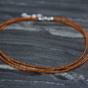 Puede incluir: Un delicado collar de varios hilos hecho de peque&ntilde;as cuentas de color marr&oacute;n anaranjado. El collar est&aacute; sobre una superficie gris oscura.