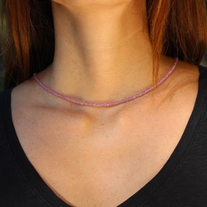 May include: A delicate pink gemstone necklace with small, faceted beads. The necklace is worn by a person with long brown hair and a black shirt.