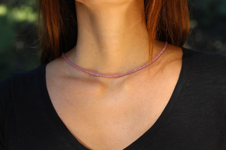 May include: A delicate pink gemstone necklace with small, faceted beads. The necklace is worn by a person with long brown hair and a black shirt.