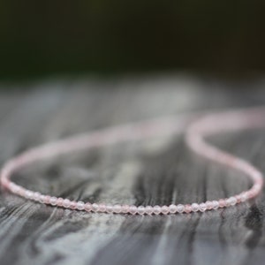May include: A delicate pink rose quartz necklace with small, round beads. The necklace is lying on a wooden surface.
