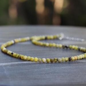 Pode incluir: Um colar delicado de pedras preciosas verdes e amarelas com um fecho de prata. O colar é feito de pequenas contas redondas e está sobre uma superfície cinza.