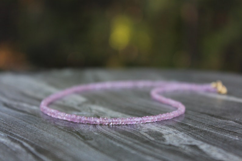 May include: A delicate pink gemstone necklace with a gold clasp. The necklace is made of small, faceted gemstones that are strung together on a thin chain.