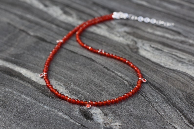 Puede incluir: Un collar de piedras preciosas de cornalina roja con detalles plateados. El collar est&aacute; hecho de peque&ntilde;as cuentas redondas y tiene un aspecto delicado y femenino.