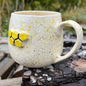 Può includere: Tazza in ceramica color crema con macchie gialle e un motivo a nido d'ape. La tazza ha una forma arrotondata e un manico curvo. Il nido d'ape è composto da esagoni gialli con contorni neri. La tazza è appoggiata su un pezzo di legno.