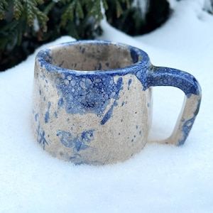 Op de afbeelding: Een handgemaakte keramische mok met een gespikkelde beige body en een donkerblauwe rand en handvat. De mok heeft blauwe spetters en sneeuwvlokachtige ontwerpen. De mok staat in de sneeuw.