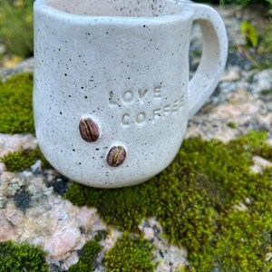 以下が含まれることがあります： 前面に「LOVE COFFEE」の文字がエンボス加工された、斑点のある白いセラミックマグ。テキストの下には2つのコーヒー豆の装飾があります。マグカップは湾曲したハンドルを持ち、苔で覆われた岩の上に置かれています。