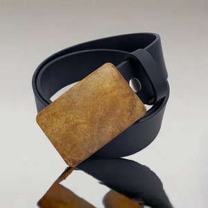 Puede incluir: Un cinturón de cuero negro con una hebilla de metal dorado. La hebilla tiene una superficie texturizada.