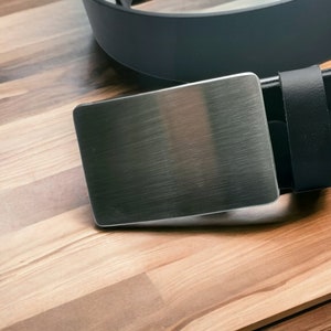 Puede incluir: Una hebilla de cinturón de metal plateado con acabado cepillado. La hebilla está unida a un cinturón de cuero negro.