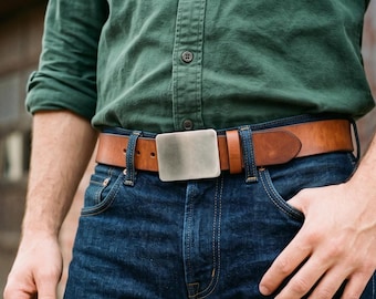 May include: A tan leather belt featuring a smooth, rectangular, brushed metal buckle. The belt is worn through the belt loops of dark blue denim jeans, positioned below a dark green button-down shirt with rolled-up sleeves.