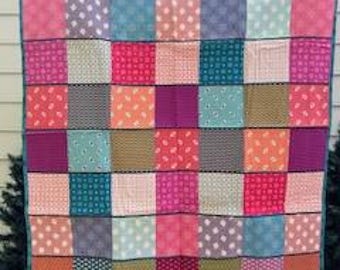 Puede incluir: Un edredón de retazos colorido con una variedad de cuadrados estampados en tonos de rosa, naranja, turquesa, morado y verde. El edredón está bordeado con tela turquesa y parece hecho a mano.