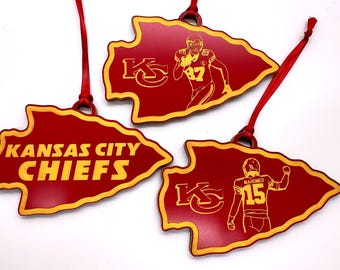 Op de afbeelding: Drie rode en gouden Kansas City Chiefs ornamenten in een pijlvorm. Eén ornament heeft de tekst "KANSAS CITY CHIEFS", de andere twee hebben illustraties van spelers en het teamlogo. Elk ornament heeft een rood lint om op te hangen.