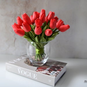 May include: A bouquet of red artificial tulips in a clear glass vase with a green ribbon. The vase is sitting on top of a book with the word "VOGUE" printed on the cover.