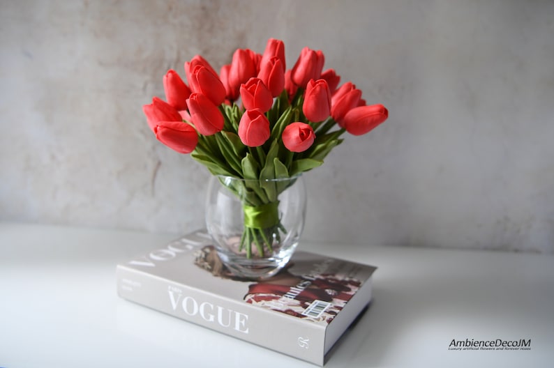 May include: A bouquet of red artificial tulips in a clear glass vase with a green ribbon. The vase is sitting on top of a book with the word "VOGUE" printed on the cover.
