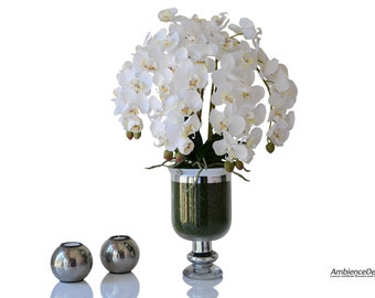 May include: A white orchid arrangement in a silver and green vase. The vase has a silver rim and a green base. Two silver candle holders are in the foreground.