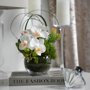 May include: White orchid flower arrangement in a clear glass vase with green moss and greenery. The vase is sitting on a book titled "The Fashion Book".