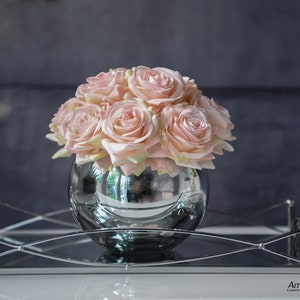 Op de afbeelding: Een boeket roze blushrozen gerangschikt in een zilveren spiegelvaas. De vaas staat op een zilveren spiegelblad.