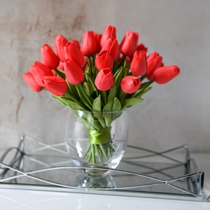 May include: A bouquet of red artificial tulips in a clear glass vase with a green ribbon tied around the stems. The vase is sitting on a silver tray with a mirrored surface.