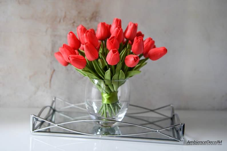 May include: A bouquet of red artificial tulips in a clear glass vase with a green ribbon tied around the stems. The vase is sitting on a silver tray with a mirrored surface.