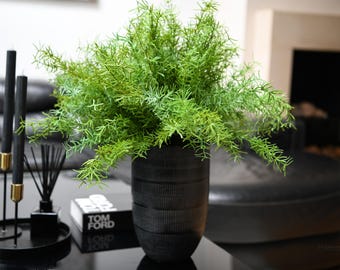 以下が含まれることがあります： 黒のテクスチャのある鉢に、細長い葉を持つ緑色の造花が植えられています。植物は、黒い本と黒い燭台のある黒いテーブルの上に置かれています。