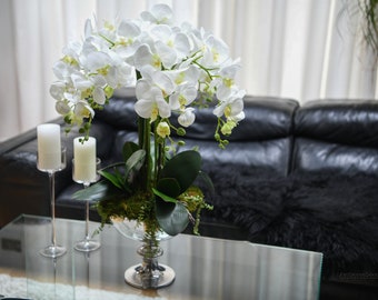 May include: A white orchid arrangement in a clear glass vase with a silver base. The arrangement is on a glass coffee table in front of a black leather sofa.