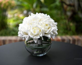 May include: A bouquet of white roses arranged in a clear glass vase. The roses are in a round shape and are sitting on a black table.
