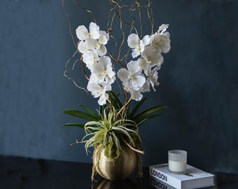 May include: A floral arrangement featuring white orchids, green leaves, and golden branches in a gold-colored, round pot. A small candle and a book with the word "LONDON" are also visible.