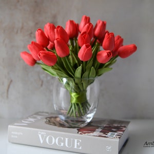 May include: A bouquet of red artificial tulips in a clear glass vase. The vase is sitting on top of a book with the word "VOGUE" printed on the cover.