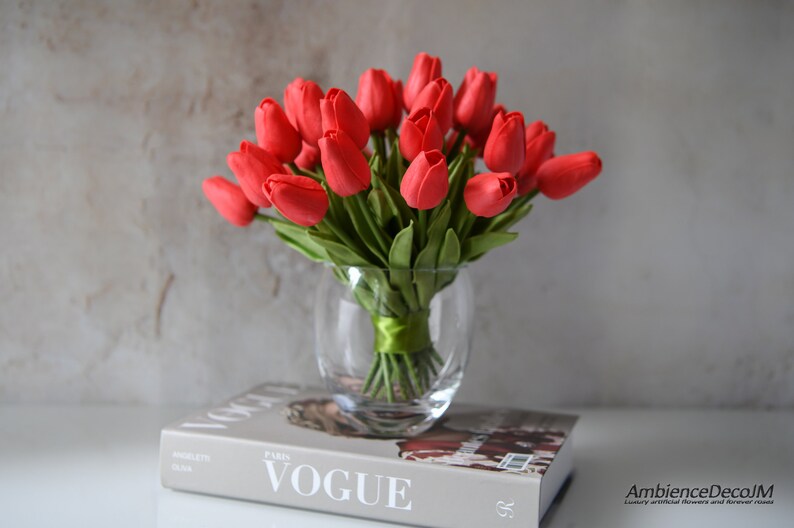 May include: A bouquet of red artificial tulips in a clear glass vase. The vase is sitting on top of a book with the word "VOGUE" printed on the cover.