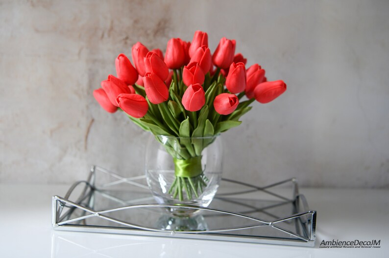 May include: A bouquet of red artificial tulips in a clear glass vase with a green ribbon tied around the stems. The vase is sitting on a silver tray with a mirrored surface.