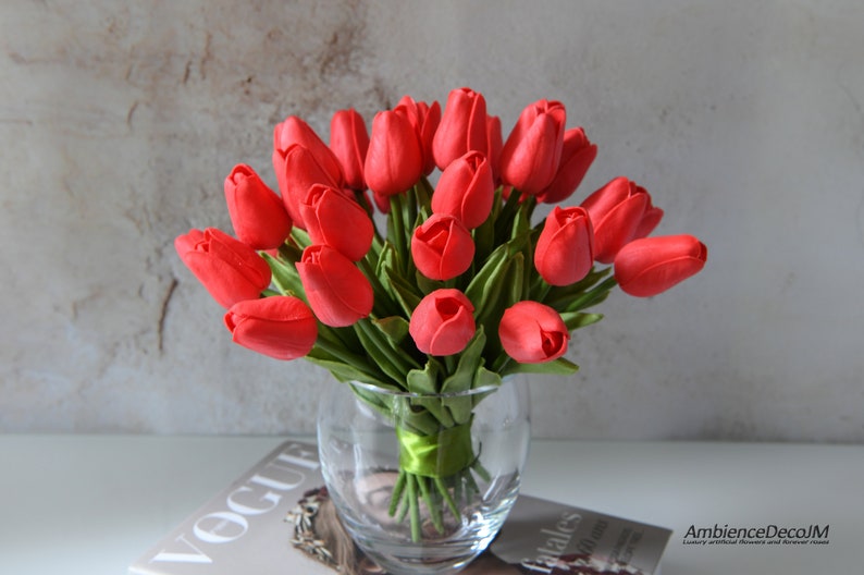 May include: A bouquet of red tulips arranged in a clear glass vase. The vase is sitting on a magazine with the word "Vogue" visible.